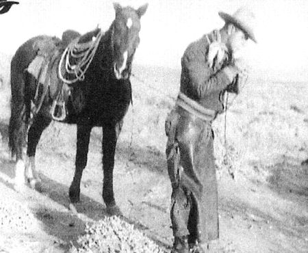 A cowboy stands next to a saddled horse on a dirt path, looking down and adjusting something on his shoulder.