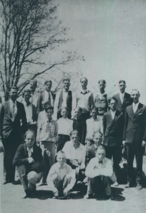 A black and white photo of twenty men posing outdoors in two rows, with trees visible in the background.