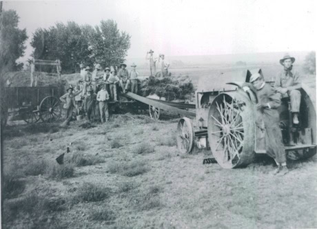 Black and white photo of several people using early 20th-century farming machinery to harvest hay, with a tractor and wagon visible in a rural field.