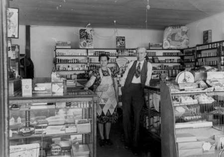 Two people stand behind the counter of a small, vintage grocery store, surrounded by shelves stocked with canned goods and other products.