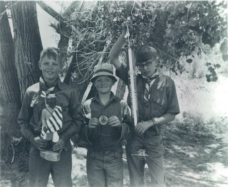 Three boys in scout uniforms stand outdoors by trees; one holds a trophy and flag, another displays a wire loop, and the third holds a large object, possibly a fishing pole.