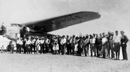 A large group of people stands in front of a vintage airplane on a dirt airstrip under a clear sky.