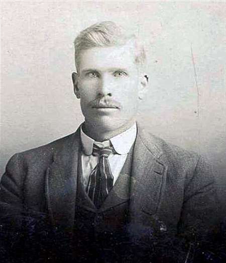 Black and white portrait of a man with short light-colored hair and a mustache, wearing a suit, vest, collared shirt, and patterned tie, facing the camera against a plain background.