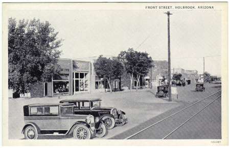 Black and white photo of vintage cars parked along a street lined with shops and trees in Holbrook, Arizona.