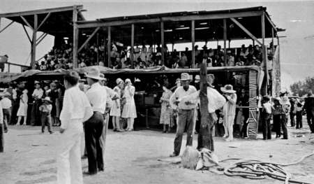 Black and white photo of a crowd gathered around and inside a two-story wooden structure, with people dressed in mid-20th-century clothing.