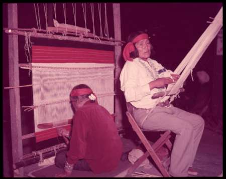 Two people wearing headbands sit at a loom, weaving a textile with red and white horizontal stripes.