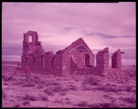 A person stands near the stone ruins of a partially collapsed building in a barren, open landscape under a cloudy sky.