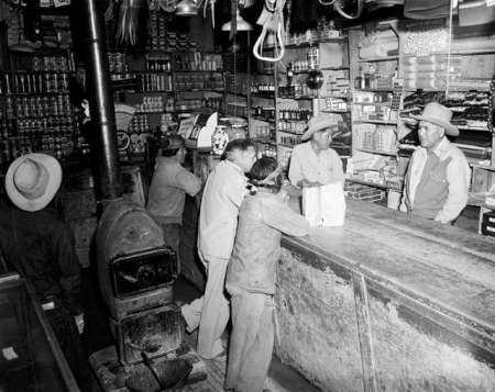 Black-and-white photo of several men in hats standing at the counter of a small, rustic general store filled with goods and supplies.