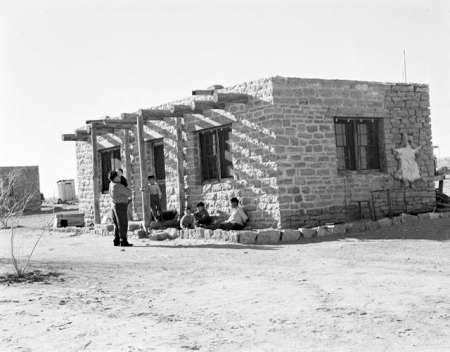 A group of people sit and stand outside a small, single-story stone house in a dry, sparse landscape.