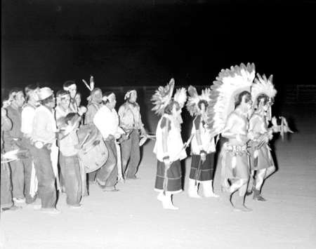 A group of people, some wearing feathered headdresses and traditional attire, stand and walk together at night on an open ground.