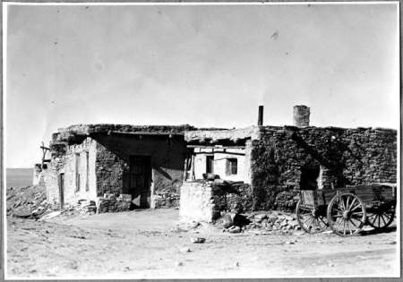 An old adobe house with a flat roof stands in a barren landscape; a wooden wagon is parked to the right of the building.