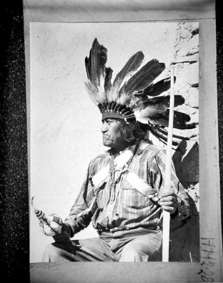 A Native American man in traditional attire, wearing a feathered headdress and holding a staff and an object, sits against a stone wall.