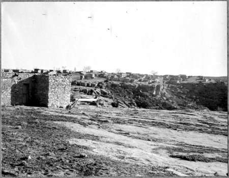 A distant view of a flat, rocky landscape with stone structures and low buildings spread across the horizon under a clear sky.