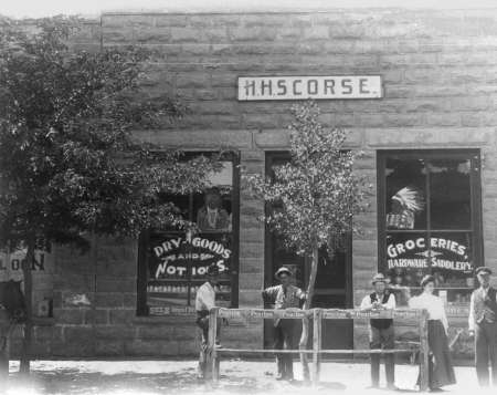 Black-and-white photo of people standing in front of an old stone building with signs reading "H.H. Scorse," "Dry Goods," and "Groceries, Hardware, Saddlery.