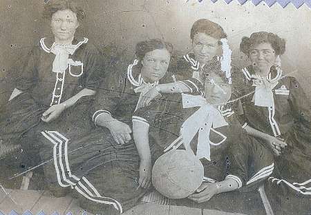 Four women in old-fashioned uniforms pose for a vintage photograph with a basketball, suggesting they are a historical women's basketball team.