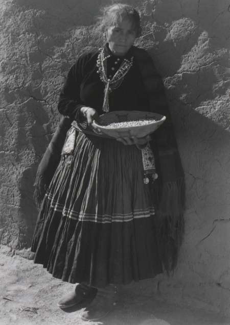 An older woman in traditional clothing stands against an adobe wall, holding a shallow bowl filled with beans or seeds.