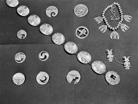 Silver jewelry pieces, including a necklace, bracelet, brooches, and pendants, displayed on a dark fabric background. Designs feature geometric and animal motifs.