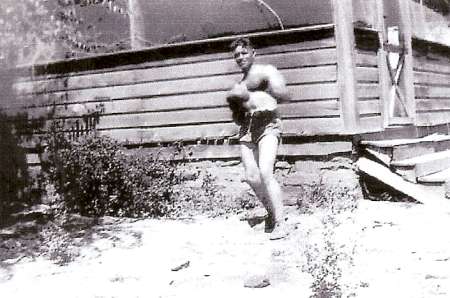 A boy in swim trunks stands outdoors holding a small animal, possibly a cat, in front of a wooden building with steps.