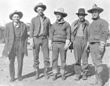Five men wearing hats and work clothes stand outdoors in a line, posing for the camera. The background is an open, rural setting.