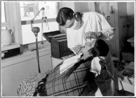 A dentist wearing a white coat examines or treats a patient who is reclining in a dental chair, with dental tools and equipment visible nearby.
