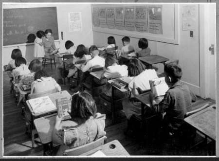 Black and white photo of a classroom with young students sitting at desks facing a teacher at the front, reading books.
