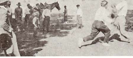 A group of people, including a horse, watches two men engaged in a fight outdoors on a dirt path.