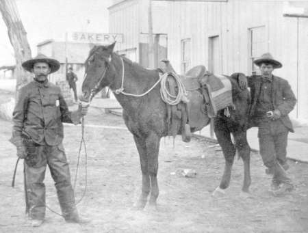 Two men in old-fashioned clothing stand with a saddled horse on a dirt street in front of wooden buildings, one labeled "Bakery.