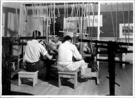 Three people sit on stools working at weaving looms in a workshop, surrounded by tools and hanging threads.