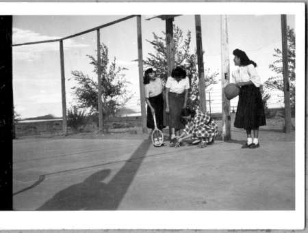 Four girls stand and kneel on an outdoor basketball court; one holds a ball, another adjusts something on the ground with a racket. Trees and fencing are visible in the background.