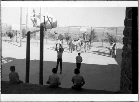 Children play basketball on an outdoor court, with several others sitting and watching from the steps in the foreground. The background features a fenced area and open landscape.