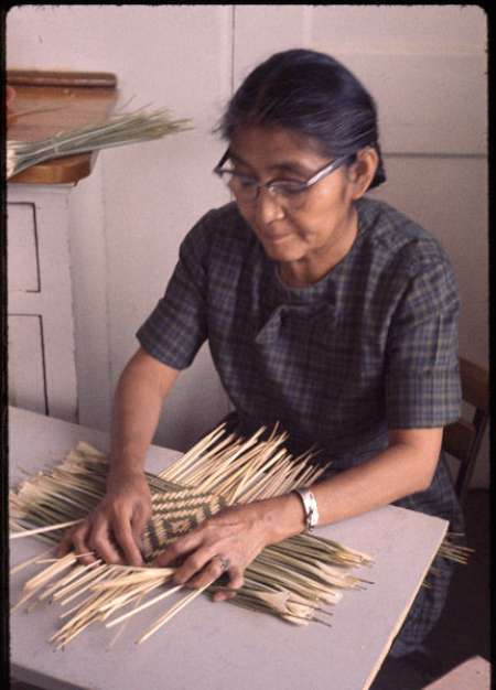 A woman with glasses weaves a basket from light-colored reeds while sitting at a table.