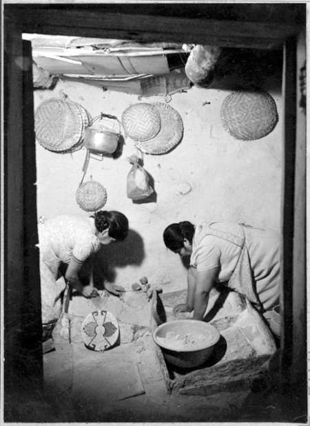 Two women kneel on the floor, preparing food in a rustic kitchen with woven baskets and utensils hanging on the wall.