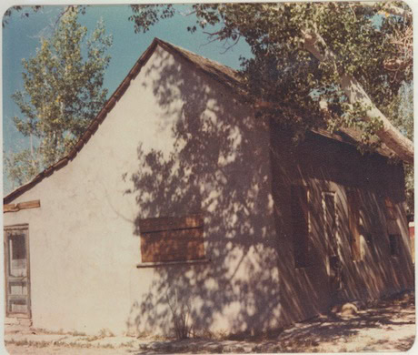 An old, small, single-story house with boarded windows and a sloped roof, partially shaded by large trees.