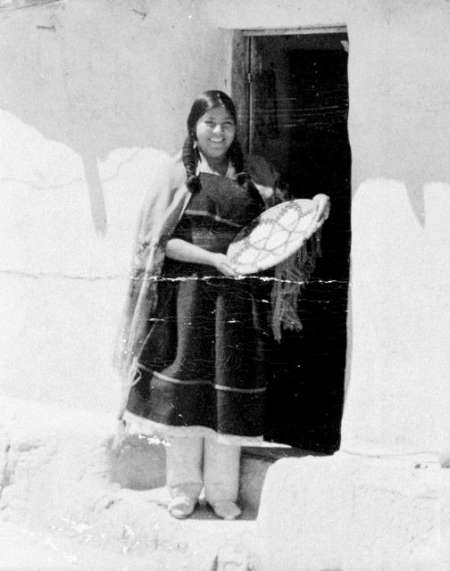 A woman stands in a doorway, wearing traditional clothing and holding a woven basket.