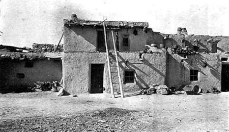 Black and white photo of an adobe two-story building with a ladder leaning to the roof and scattered debris around the base.