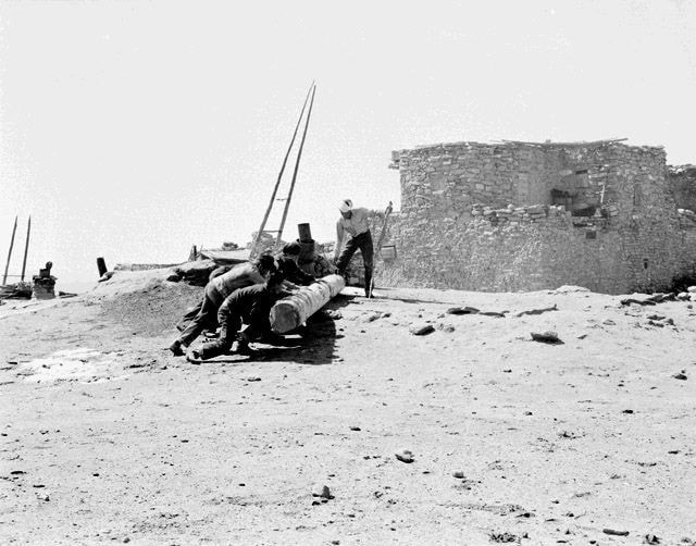 Several people push a large wooden log up a slope near a stone building in an arid, rocky landscape under a clear sky.