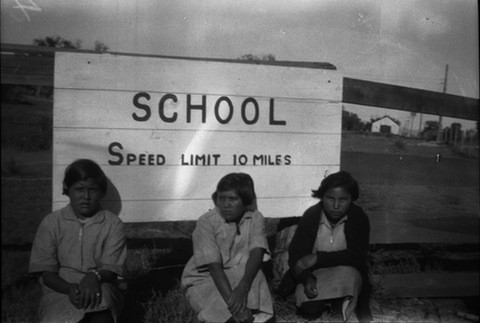 Three young girls sit in front of a large sign that reads "SCHOOL SPEED LIMIT 10 MILES," with a rural background visible behind them.