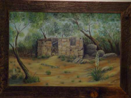 A painting of an old stone building in a wooded area, surrounded by trees, rocks, and sparse vegetation, framed with rustic wood.