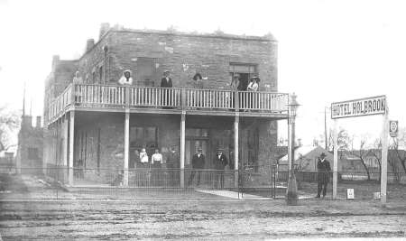 Black-and-white photo of people standing on the porch and balcony of a two-story brick building with a sign reading "Hotel Holbrook.