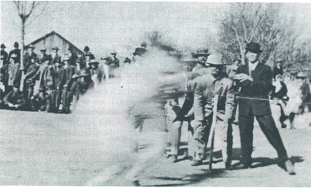 A black-and-white photograph showing a group of men watching a blurred figure running past them outdoors, with a barn and trees in the background.