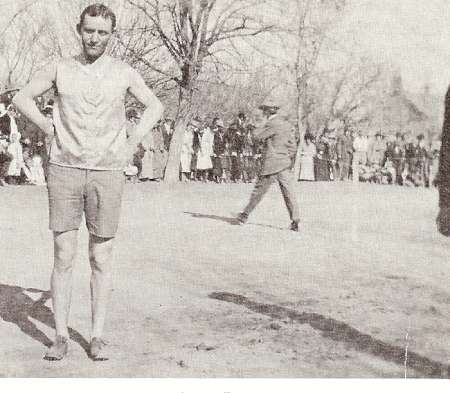 A man in athletic clothes stands outdoors on dirt, with his hands on his hips. A crowd of people and bare trees are visible in the background.