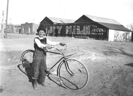 A boy in overalls stands outdoors holding a bicycle with a basket; behind him are barns with signs reading "CITY MILK" and "THE ARIZONA PRODUCE.