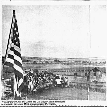 A group of people with instruments gather outdoors near an American flag, with open fields and buildings in the background.