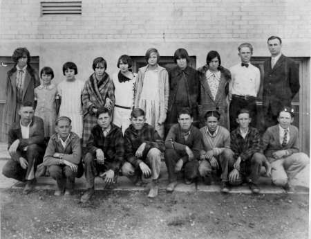 A black and white photo of a group of students and one adult posed in two rows outside in front of a brick building.