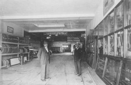 Two men stand inside a large workshop or store with shelves of tires on the left and glass display cases with tools or parts on the right.