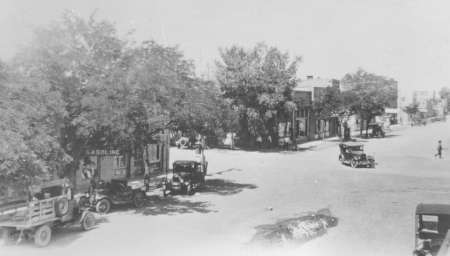 Black and white photo of a quiet street intersection with several vintage cars parked along buildings and trees lining the sidewalk.