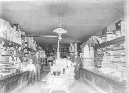 Black-and-white photo of an old-fashioned general store interior with four people standing among shelves stocked with various goods.