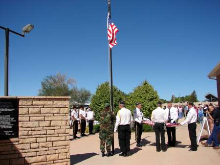 A group of people, including uniformed individuals, gather around a flagpole with the American flag raised, participating in a flag ceremony outdoors on a clear day.
