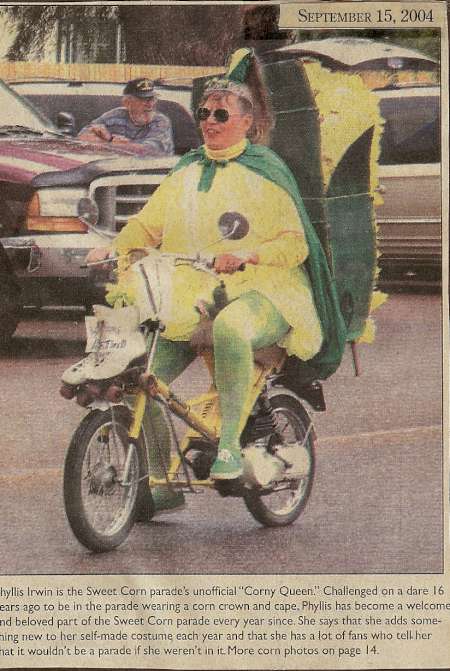 A person dressed as a corn cob, with a crown and green cape, rides a small bicycle in a parade. The newspaper clipping is dated September 15, 2004.