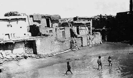 Three children walk across a muddy open area in front of old, weathered, multi-story adobe buildings under a bright sky.
